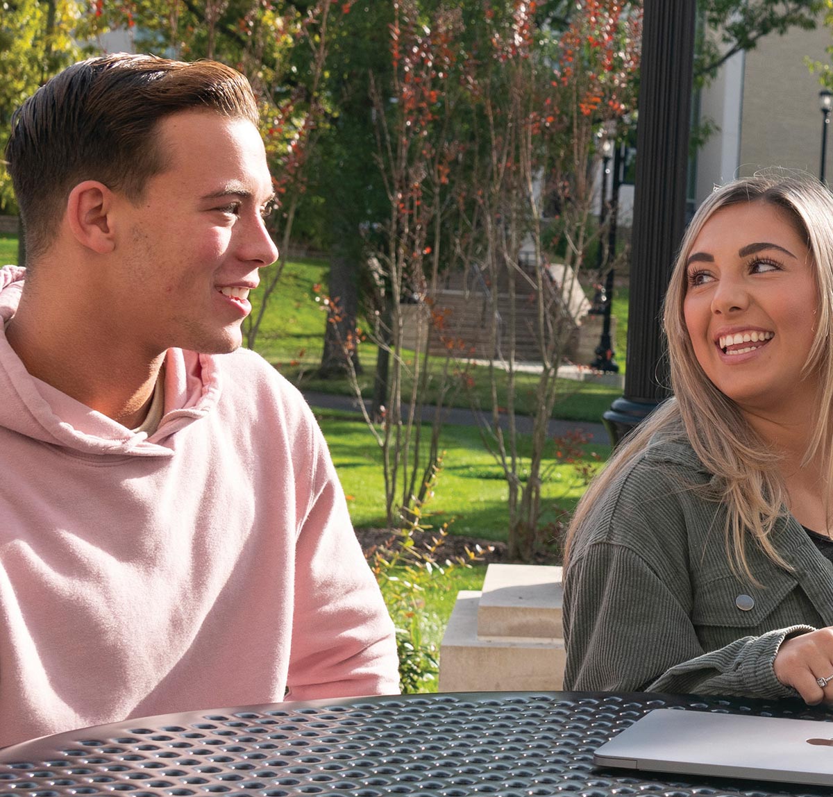 Two students sitting outside at a table as they both smile and laugh while chatting together