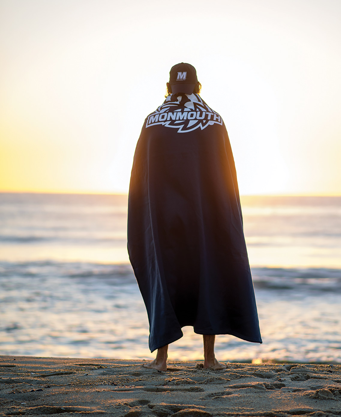 A portrait photograph of a Monmouth University student standing and posing for a picture with his beach towel over his back as he looks upon the sunset at the beach