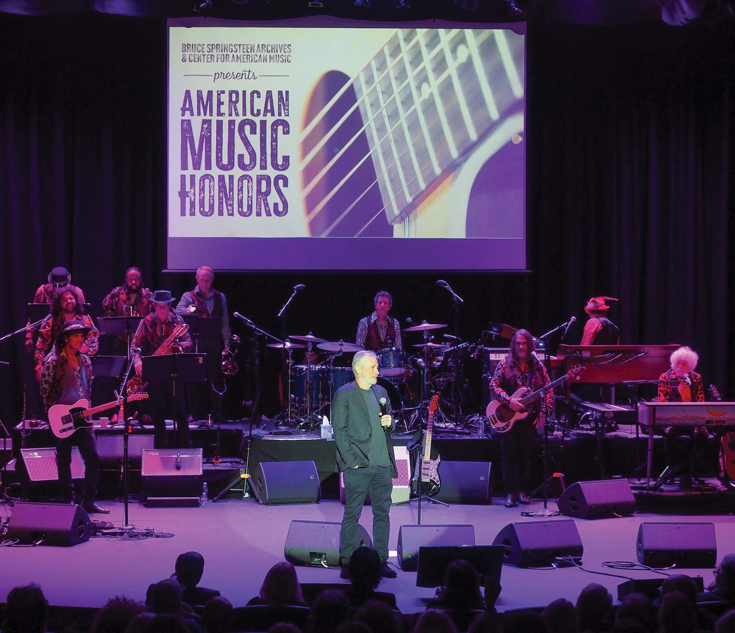 Jon Stewart in the middle center stage addressing the audience as he holds a microphone and other instrument performers behind him inside at The American Music Honors show event presented by The Bruce Springsteen Archives and Center for American Music