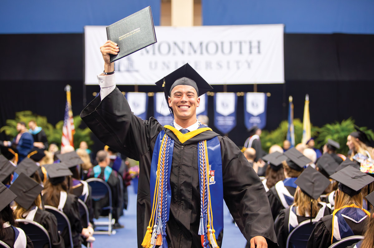 man walking down aisle at graduation ceremony while smiling at camera and holding his diploma in the air