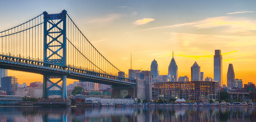 City skyline at sunset with a large suspension bridge in the foreground.