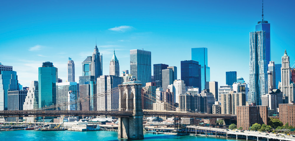 New York City skyline with the Brooklyn Bridge in the foreground under a clear blue sky.