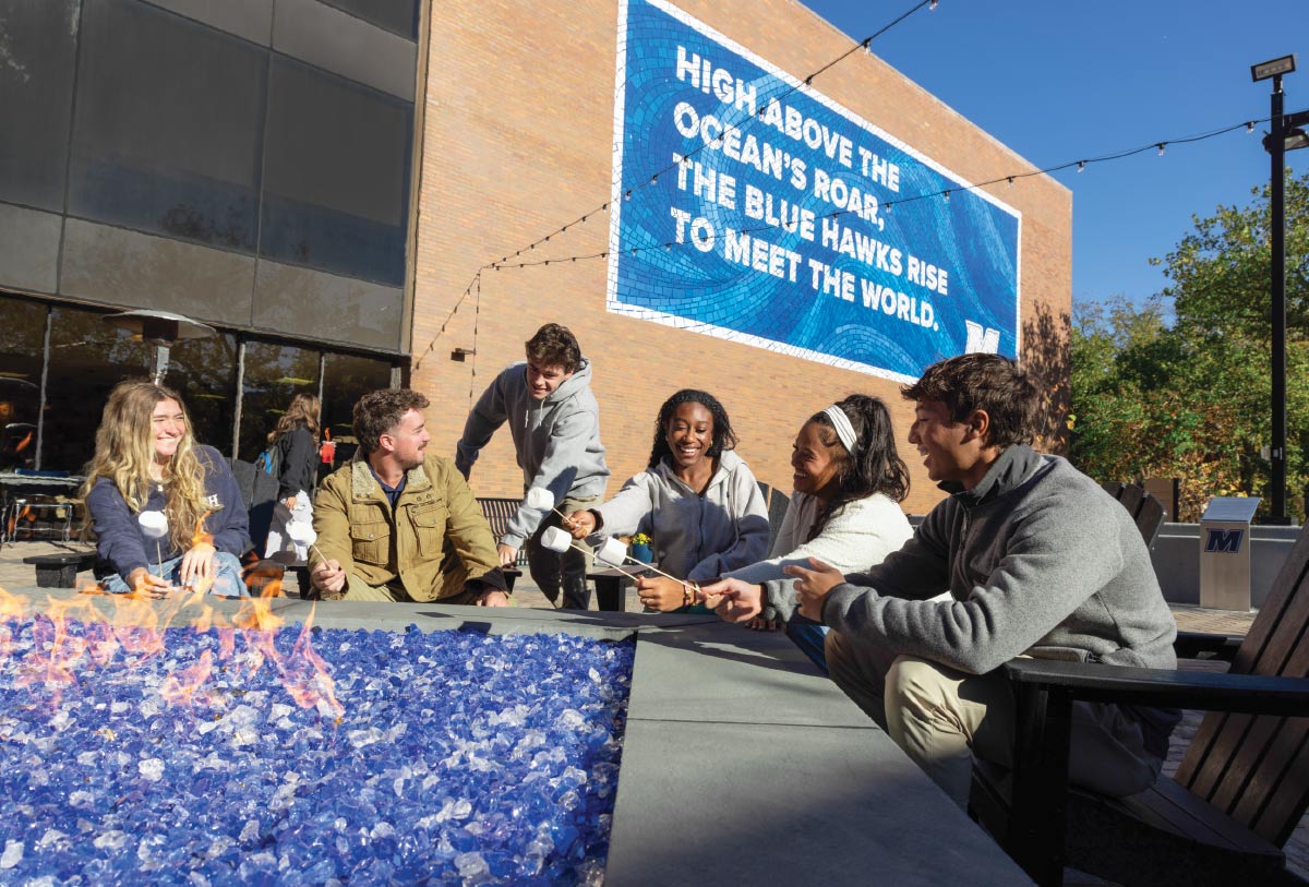 a group of six students gathered around a rectangular fire pit filled with blue glass rocks roasting marshmallows on sticks.