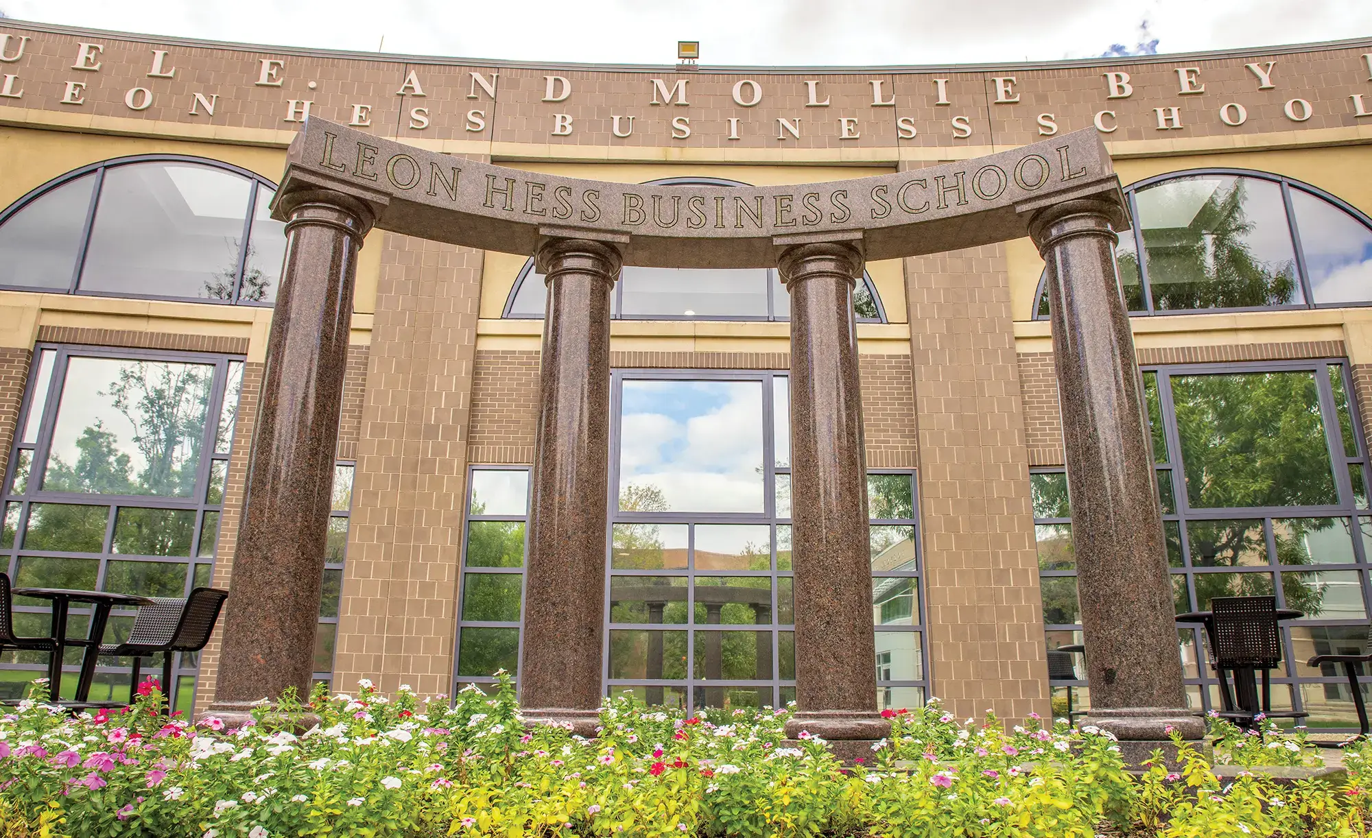 Leon Hess Business School building at Monmouth University; In the foreground, there's a vibrant flower bed filled with pink and white impatiens blooms