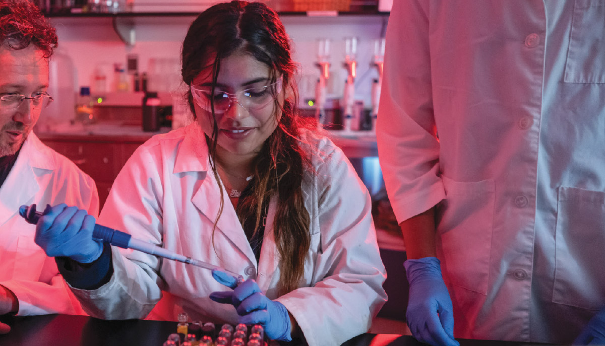 A close-up of a female student in lab coat and safety glasses using a pipette to complete an experiment under red lighting.
