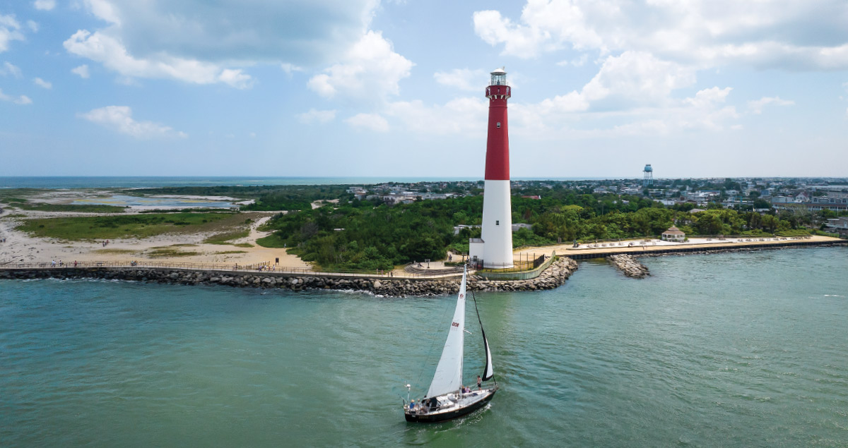 An aerial shot of a white and red lighthouse standing on a coastline, with a sailboat gliding through the water in the foreground.