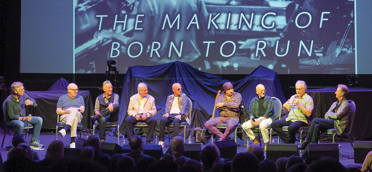 Nine men, including Bruce Springsteen, sit on stage for a panel discussion in front of a seated audience. A large screen behind them reads "THE MAKING OF BORN TO RUN."