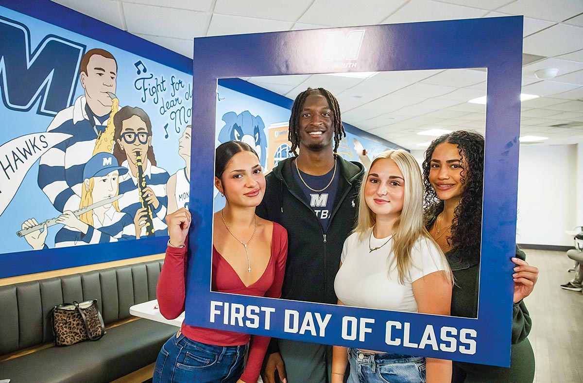 Four smiling students posing together inside a large blue photo frame that reads "FIRST DAY OF CLASS."