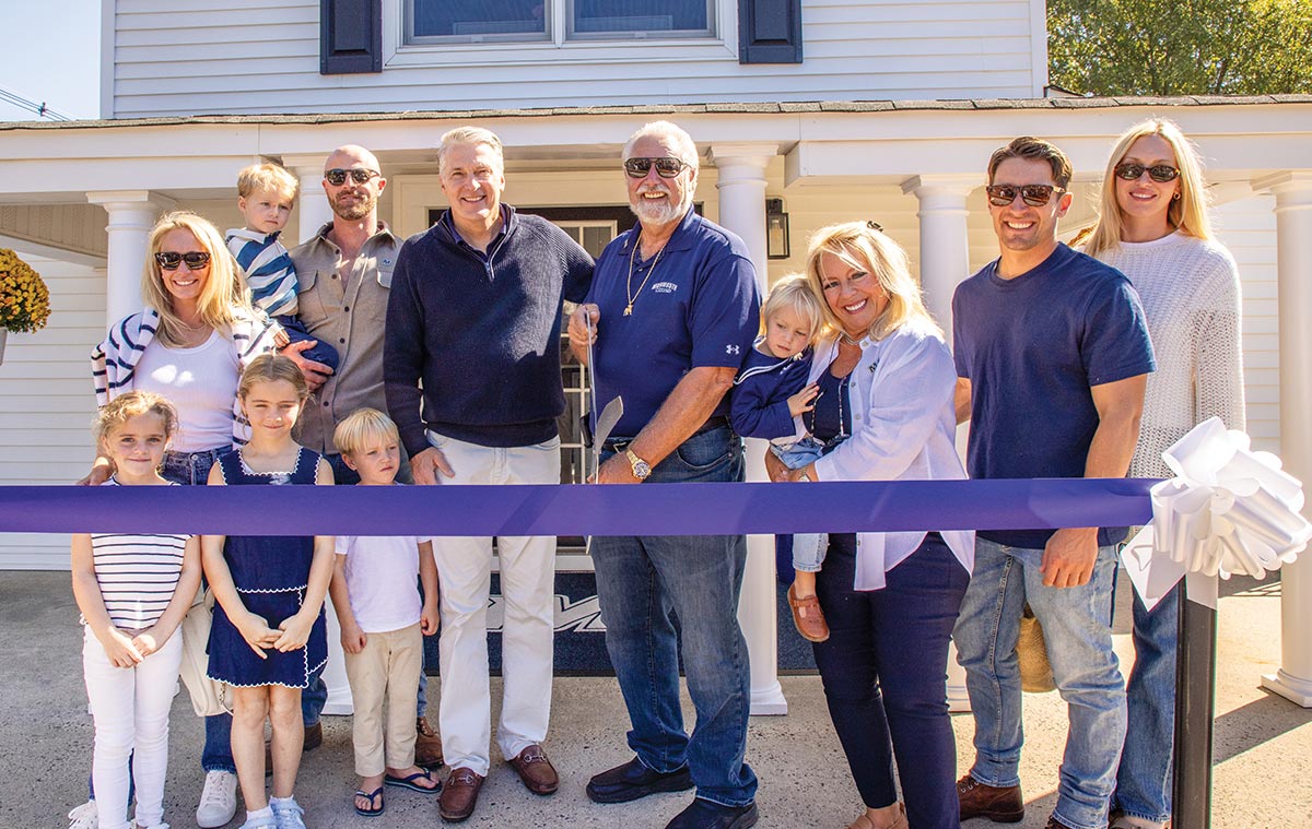 A large family group smiles together while cutting a purple ceremonial ribbon in front of a white house.