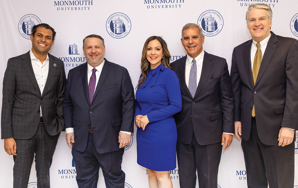 Five professionals in suits and formal wear pose together in front of a Monmouth University backdrop.