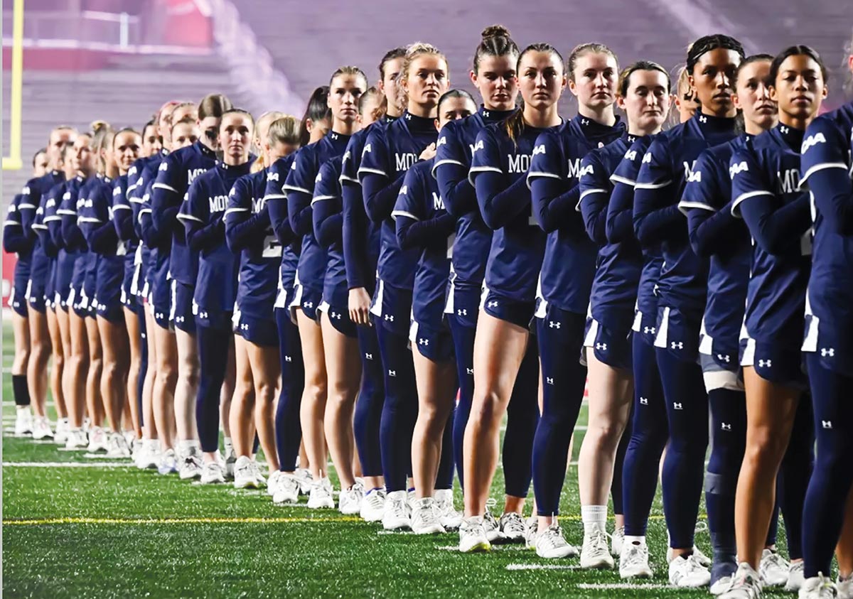 A long line of female student-athletes in matching navy uniforms stand in a row on a sports field during a ceremony.