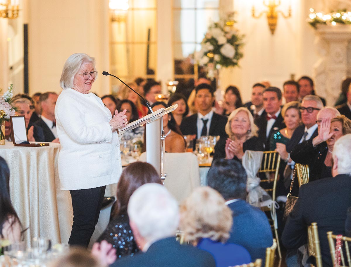 An older woman in a white blazer speaks at a podium during a formal, crowded banquet dinner.